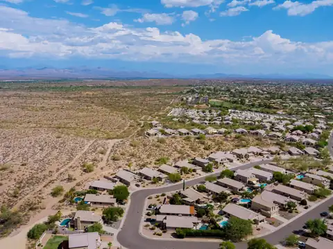 Tile-roofed houses at the edge of a desert subdivision seen from the air, with open desert beside them and mountains on the horizon