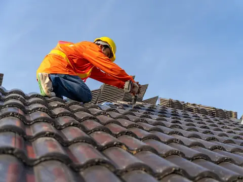 A roofer in a hard hat kneeling on a concrete tile roof and driving a screw with a cordless drill, with stacks of tiles beside him