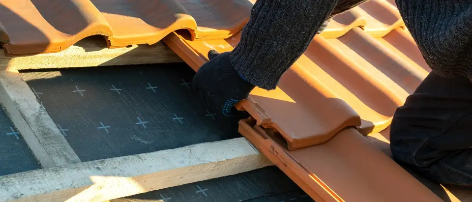 A gloved hand sliding a clay roof tile into place beside a gap that shows the wood battens and the dark underlayment beneath
