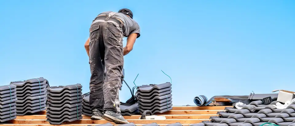 A roofer seen from behind, bending over a roof where bundles of gray tiles are stacked on the battens above the courses already laid